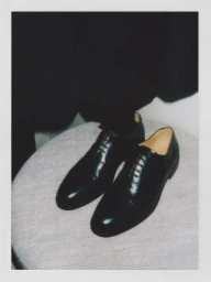 Pair of polished black leather dress shoes resting on a light grey textured round cushion in a Polaroid photo.