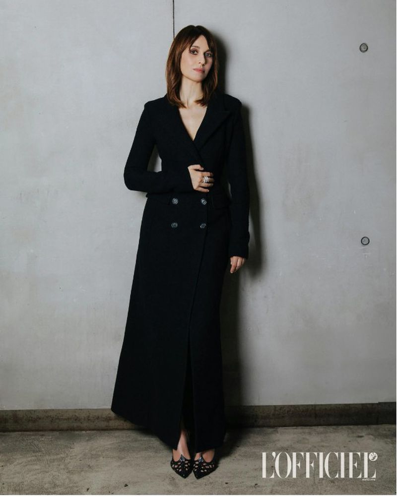 Woman in a long black coat posing against a concrete wall, fashion editorial shot, dark hair, looking at the camera, L'Officiel logo visible.