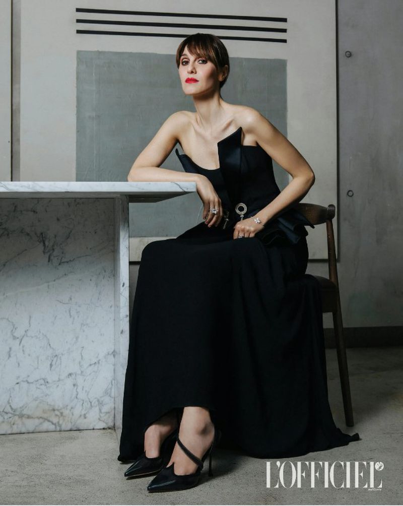 Elegant woman in a black gown sitting at a marble table, posing in front of a grey wall with black stripes and logo.