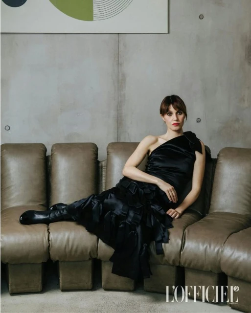 A brunette woman in a black, one-shoulder dress sits on a tan sectional sofa in an indoor space with a concrete wall and modern art.