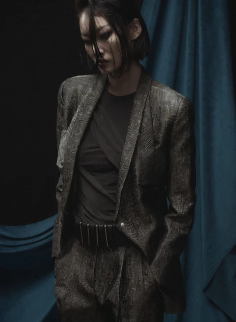 Portrait of an Asian woman in a suit, looking down against a teal curtain and dark background in a fashion or editorial photoshoot.