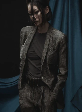 Portrait of an Asian woman in a suit, looking down against a teal curtain and dark background in a fashion or editorial photoshoot.