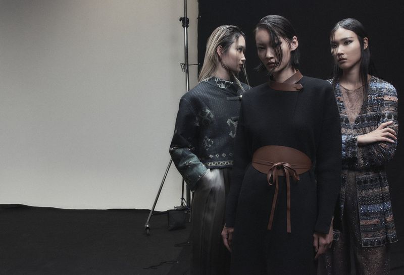 Three Asian female fashion models in a studio. The center model wears a black dress with a tan waist belt. Dark studio backdrop.