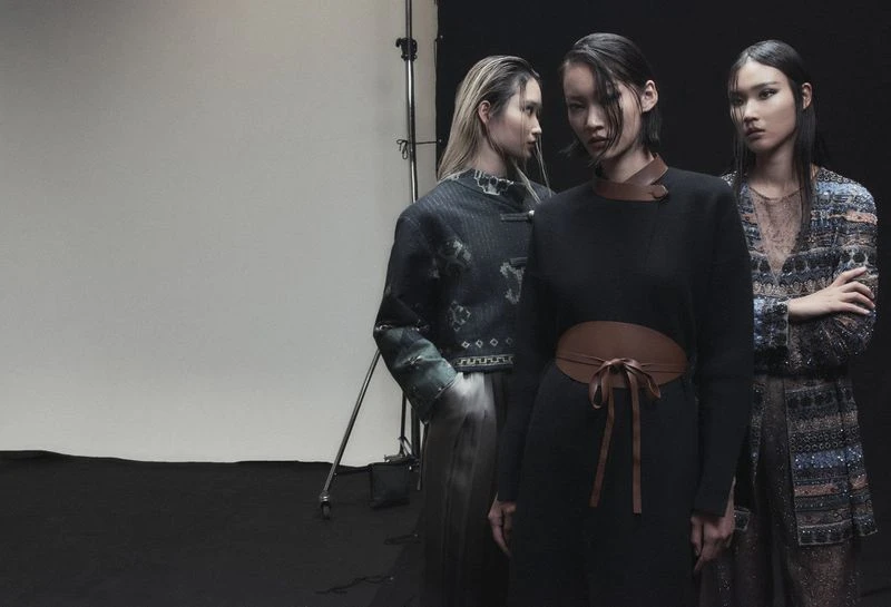 Three Asian female fashion models in a studio. The center model wears a black dress with a tan waist belt. Dark studio backdrop.