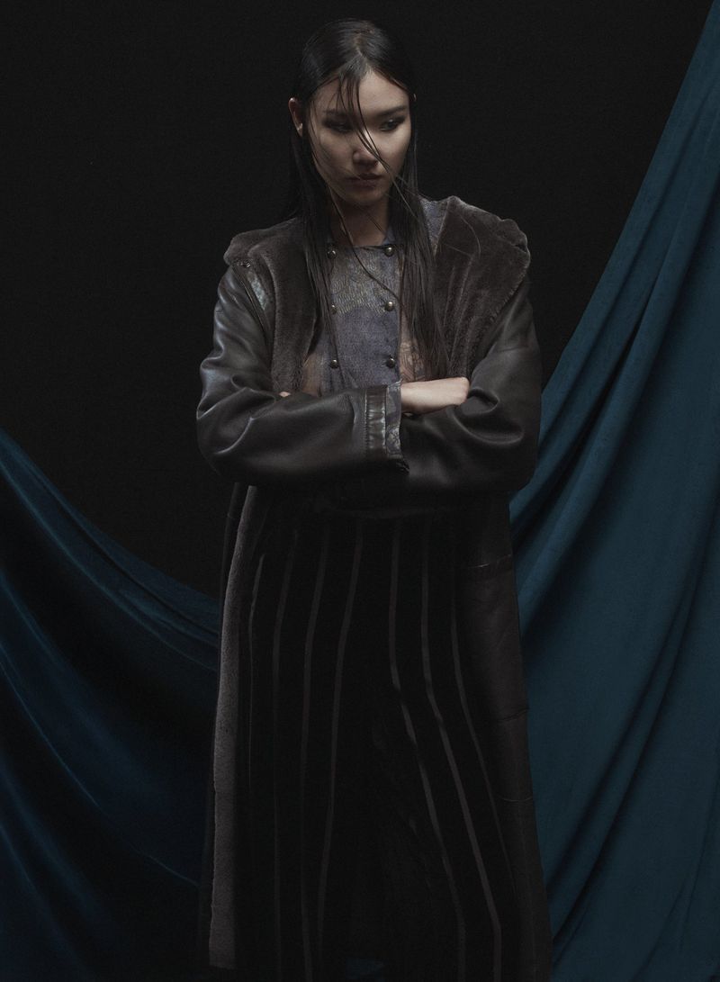 Stylish woman with dark hair in a long brown coat and pleated skirt, standing in a studio with a dark teal backdrop.