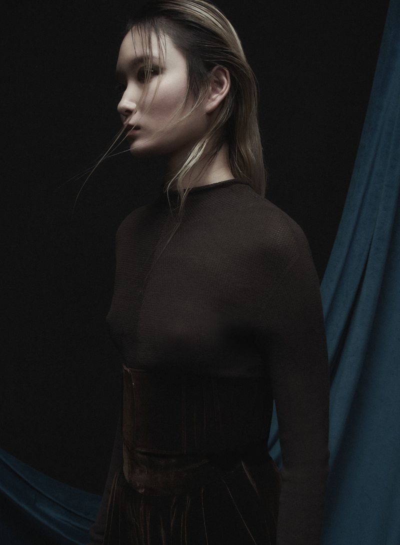 Portrait of an Asian adult female model wearing a dark, sheer top, captured in a photo studio with draped teal fabric backdrop.