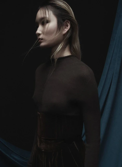 Portrait of an Asian adult female model wearing a dark, sheer top, captured in a photo studio with draped teal fabric backdrop.