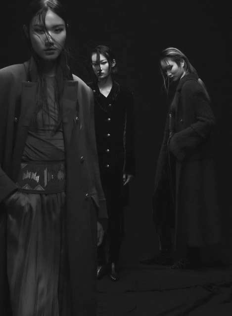 Black and white fashion shot of three Asian women in long coats posing in a dark photo studio with a spotlight on them.