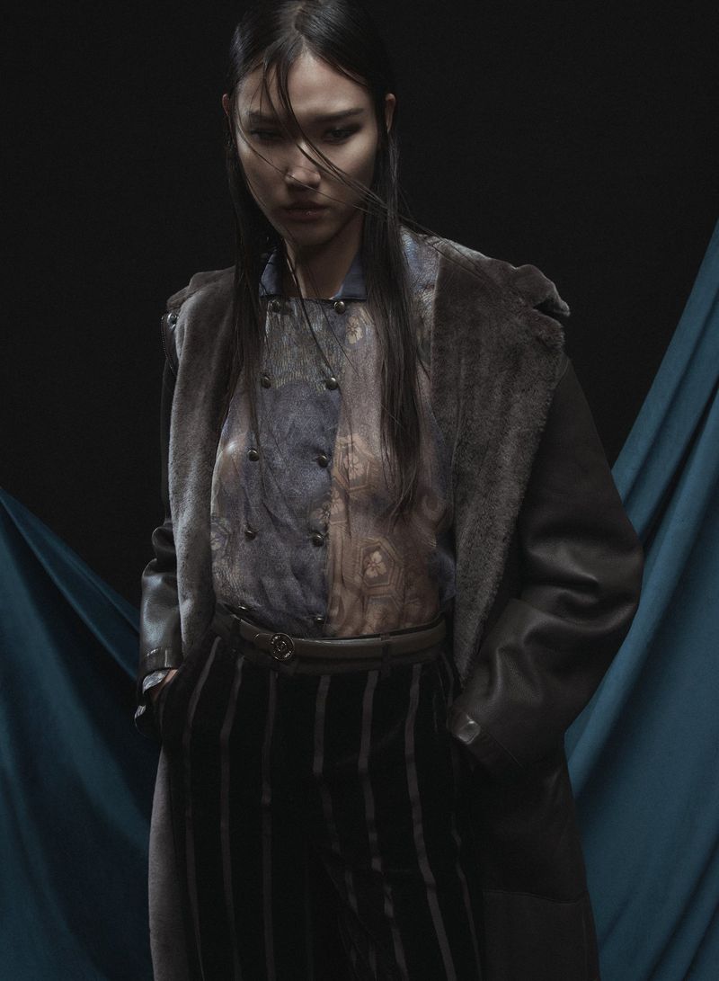A woman in an overcoat and striped pants stands against a teal and black background in a fashion-style portrait with dramatic lighting.