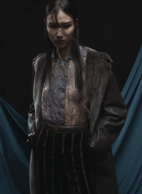 A woman in an overcoat and striped pants stands against a teal and black background in a fashion-style portrait with dramatic lighting.