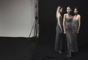 Three female models wearing grey strapless gowns standing against a dark studio backdrop with a lighting stand visible