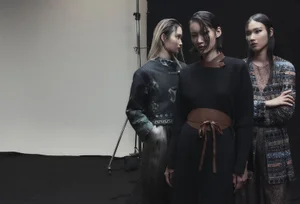 Three models posing in structured designer outfits with leather and patterned details against a studio backdrop.
