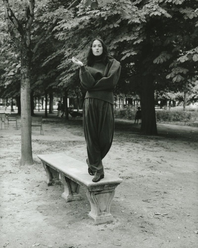Woman stands on a stone bench in a park surrounded by trees, wearing a high neck sweater and wide leg pants in a black and white photo.