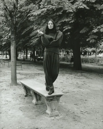 Woman stands on a stone bench in a park surrounded by trees, wearing a high neck sweater and wide leg pants in a black and white photo.