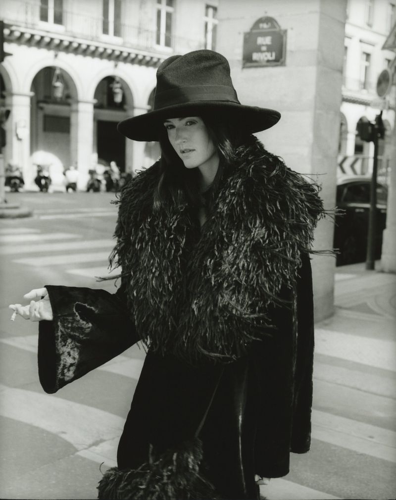 A woman in a wide-brimmed hat and large feather boa poses in a monochrome outdoor shot, with Parisian architecture in the background.