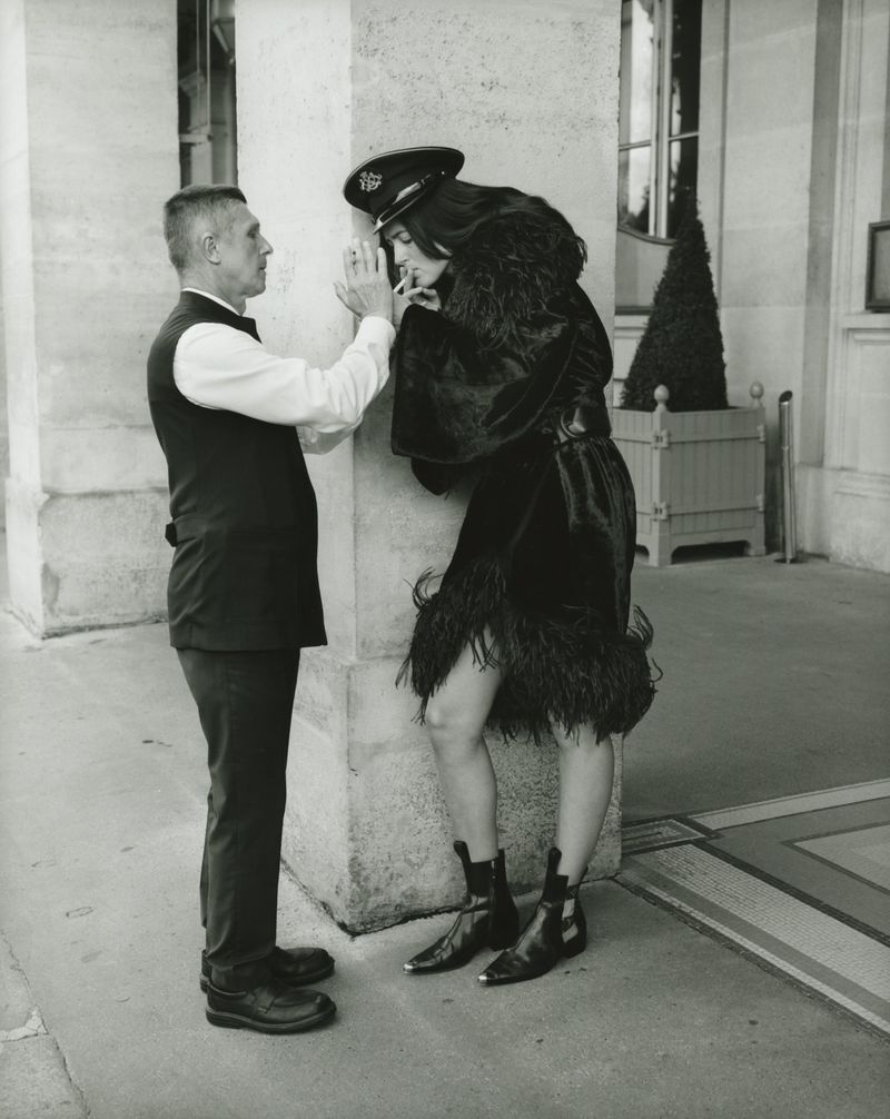 A black and white photograph shows a woman in a black outfit and cap smoking with a man standing next to a concrete pillar outdoors.