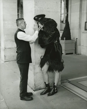 Man lighting a cigarette for a woman in a black feathered coat leaning against a stone pillar in a portico