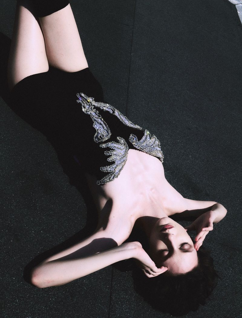 Young woman with dark hair and pale skin poses while laying on a dark surface, wearing a black dress with silver embellishments.