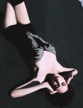 Young woman with dark hair and pale skin poses while laying on a dark surface, wearing a black dress with silver embellishments.