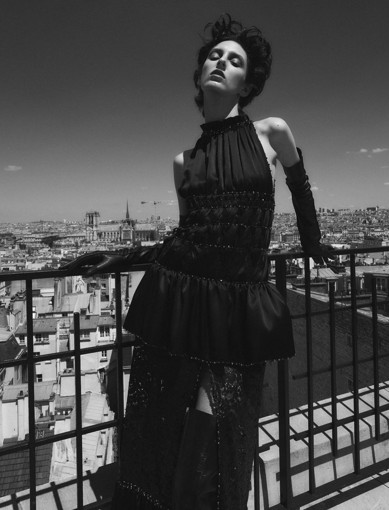 An adult woman poses on a rooftop wearing a formal black gown, opera gloves, and dark makeup, with a city skyline in the background.