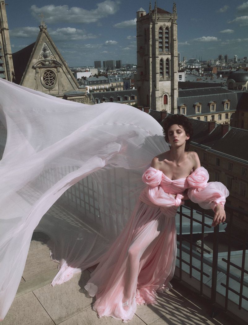 An adult woman stands on a balcony in a light pink dress with a flowing train and ornate architectural features in the background.