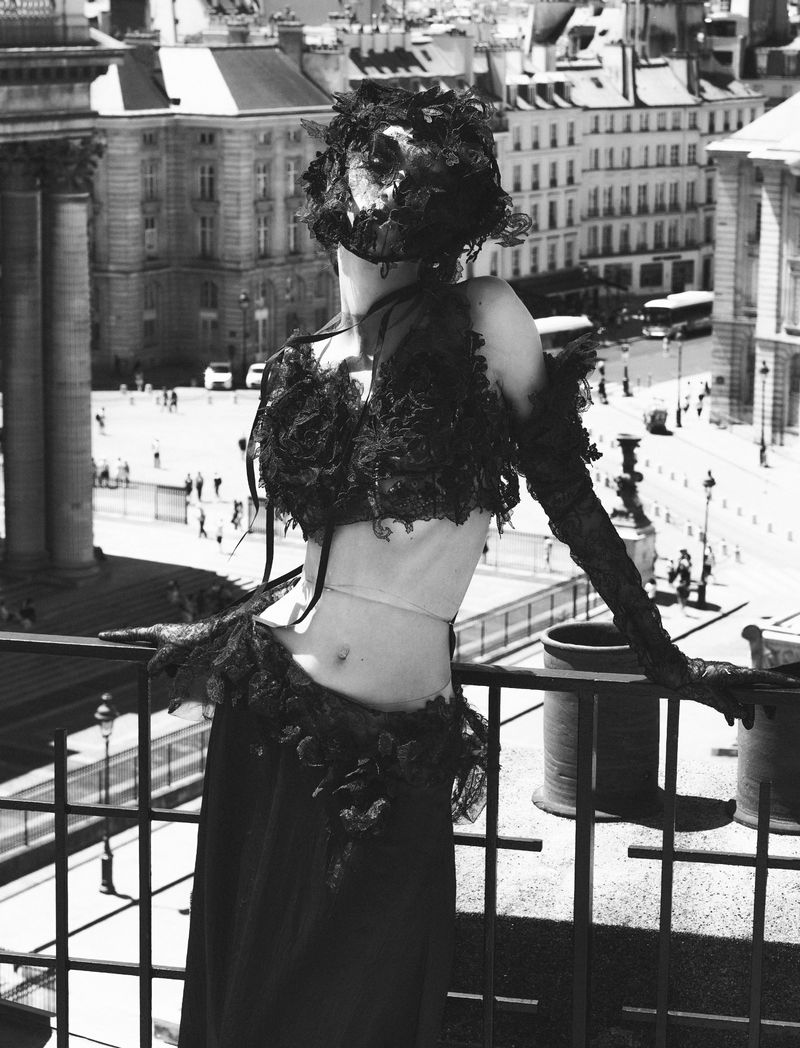 Woman wearing an elaborate lace garment poses on a balcony overlooking a city square with classical buildings in a black and white photograph.