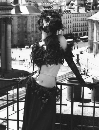 Woman wearing an elaborate lace garment poses on a balcony overlooking a city square with classical buildings in a black and white photograph.