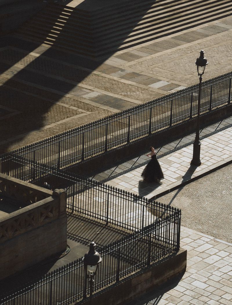 Woman in a black dress walks along a cobblestone walkway with ornate fencing and a lamp post, illuminated by shadows from nearby stairs.