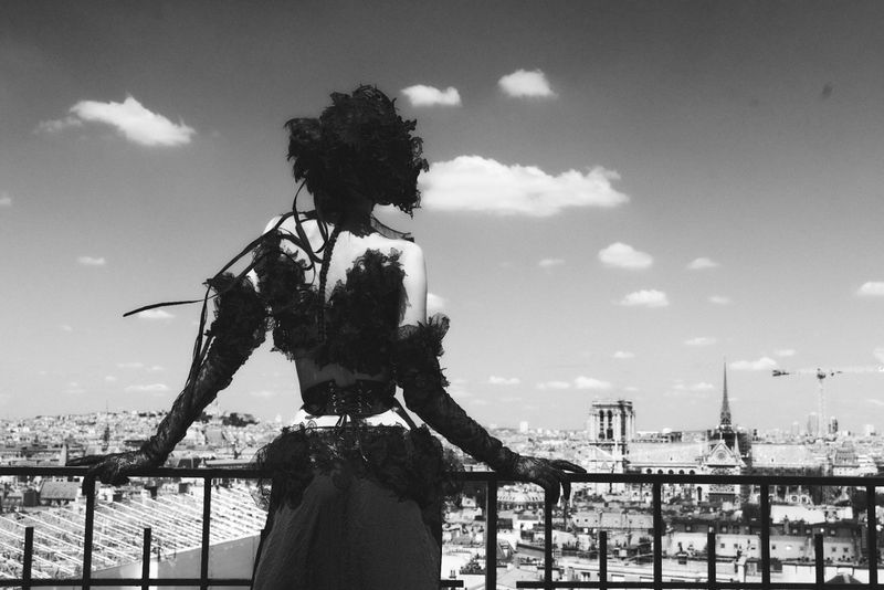 A woman in a black lace dress with gloves stands on a rooftop overlooking a cityscape with a bright sky and scattered clouds.
