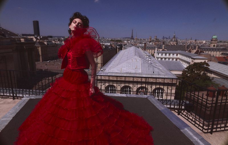 A woman in a voluminous red gown stands on a rooftop in Paris, France, with a clear sky and city buildings behind her.