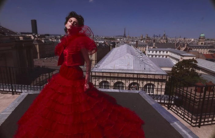 A woman in a voluminous red gown stands on a rooftop in Paris, France, with a clear sky and city buildings behind her.