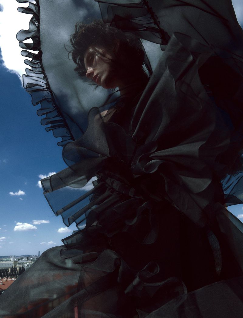 Eye-level shot of adult woman posing outdoors in black frilled dress against blue sky and clouds with urban landscape visible below.