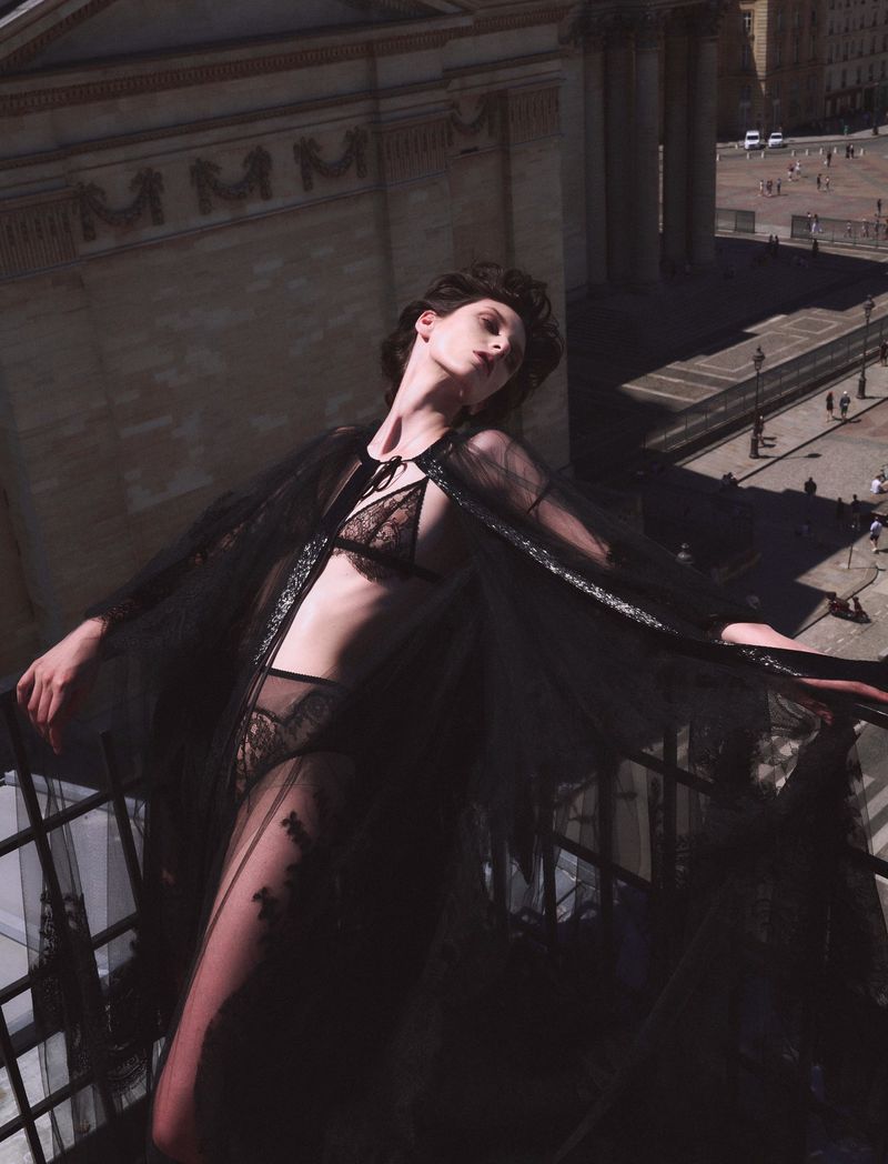 A model on a balcony in black lingerie and a sheer robe overlooks a city square with a large classical building on a sunny day.