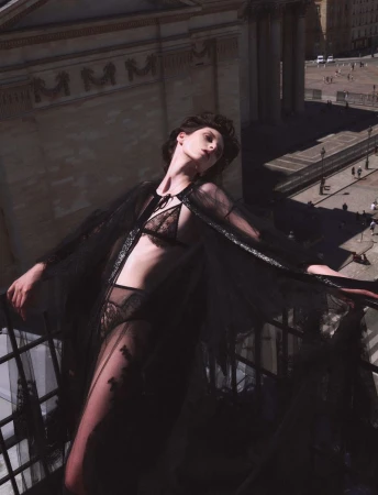 A model on a balcony in black lingerie and a sheer robe overlooks a city square with a large classical building on a sunny day.