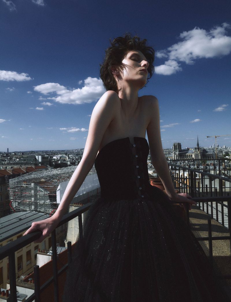 Woman in a black dress poses on a rooftop with a city skyline and blue sky with white clouds in the background during daytime.
