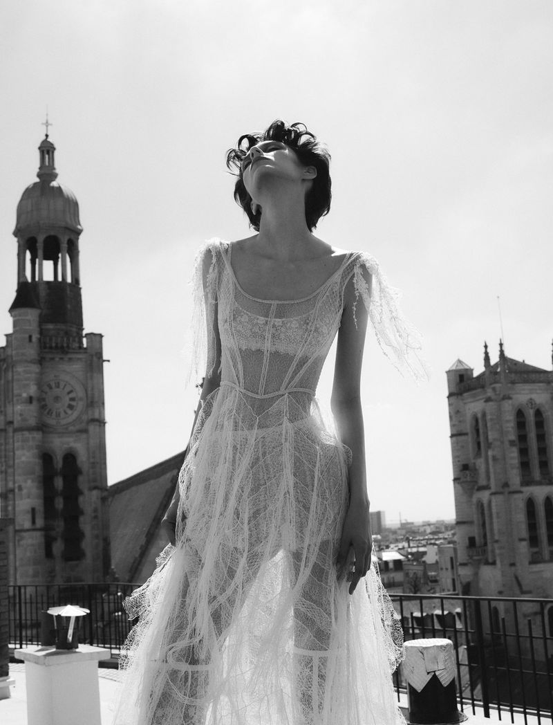 Elegant woman in a lace dress stands on a rooftop with historic buildings in the background, captured in black and white with a low angle.