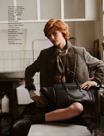 Fashion editorial shot of a young adult model with reddish-brown hair, wearing a blazer, scarf, and holding a black handbag.