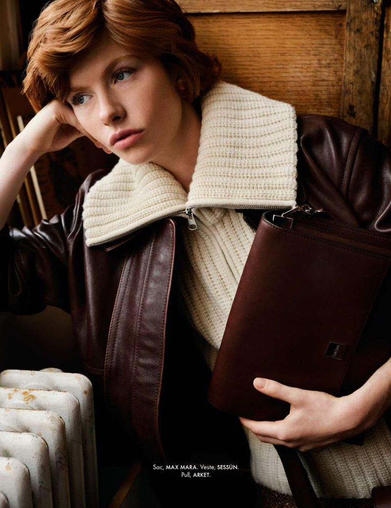 Woman in leather jacket holds a brown leather bag. Portrait-style fashion shot with a posed model indoors near white radiator.