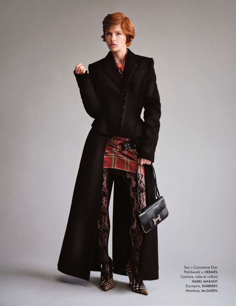 A woman with red hair stands in a studio wearing a long black coat, plaid kilt, black lace tights, and a black leather purse.