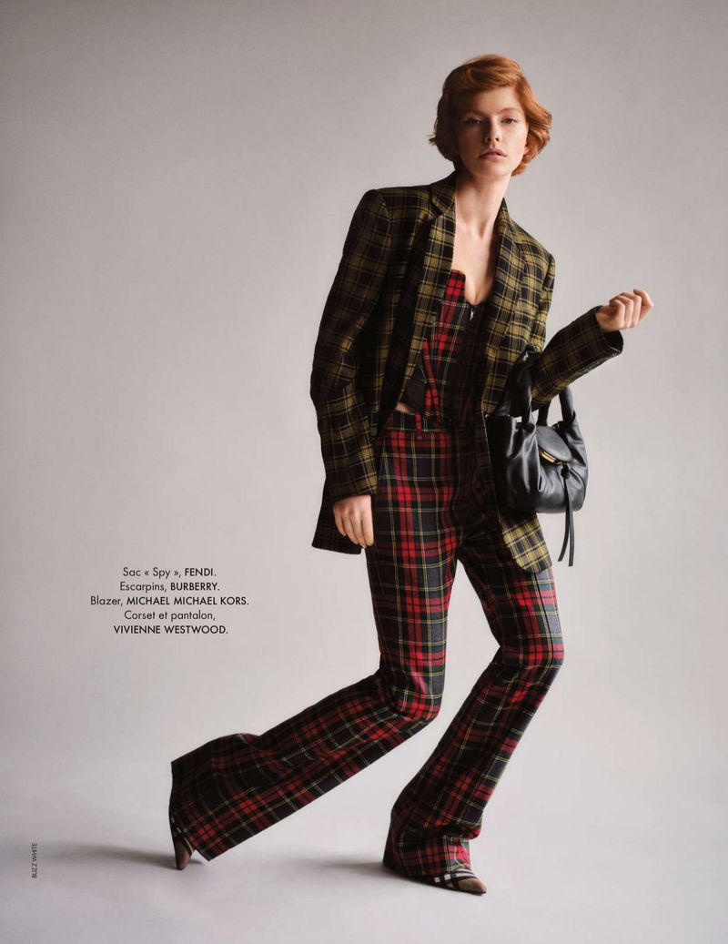 A model poses in a plaid blazer, corset, and flare pants with a black purse in a studio setting, showcasing a fashion-forward outfit.