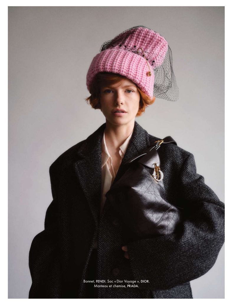 A woman in a grey coat and pink hat carries a black purse in a fashion editorial style portrait against a plain background.