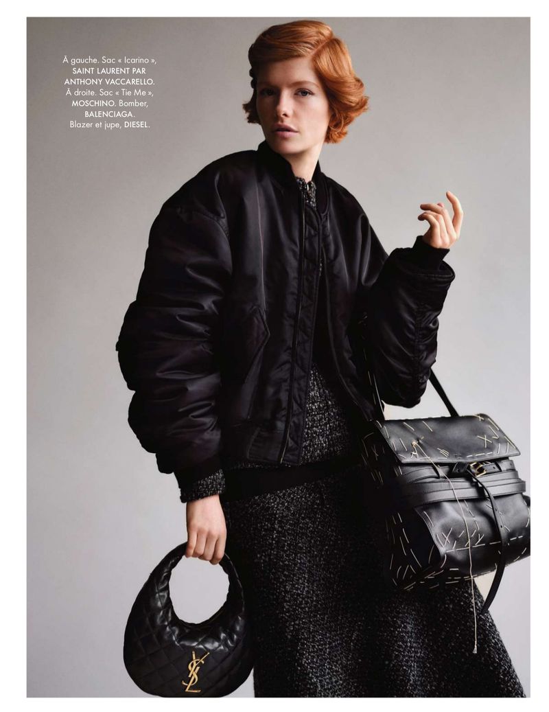 Editorial shot of a woman in a black bomber jacket holding two black leather purses. The studio background is neutral grey.