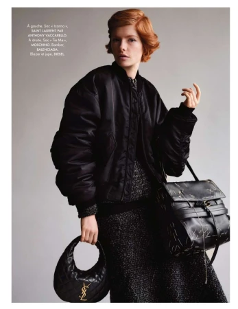 Editorial shot of a woman in a black bomber jacket holding two black leather purses. The studio background is neutral grey.