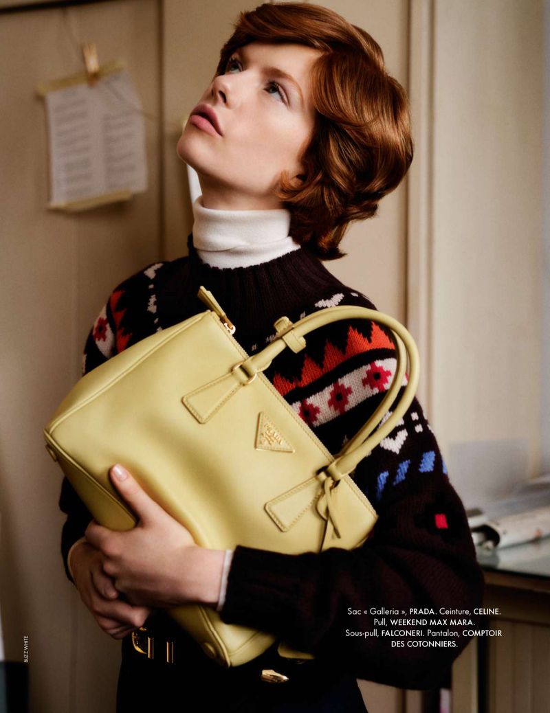 A woman with short red hair holds a yellow Prada Galleria handbag in a studio setting, wearing a patterned sweater and white turtleneck.