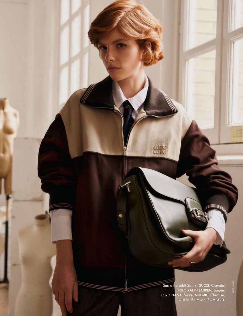 A young woman poses indoors wearing a brown and beige jacket, white collared shirt, and holding a large dark green leather satchel.