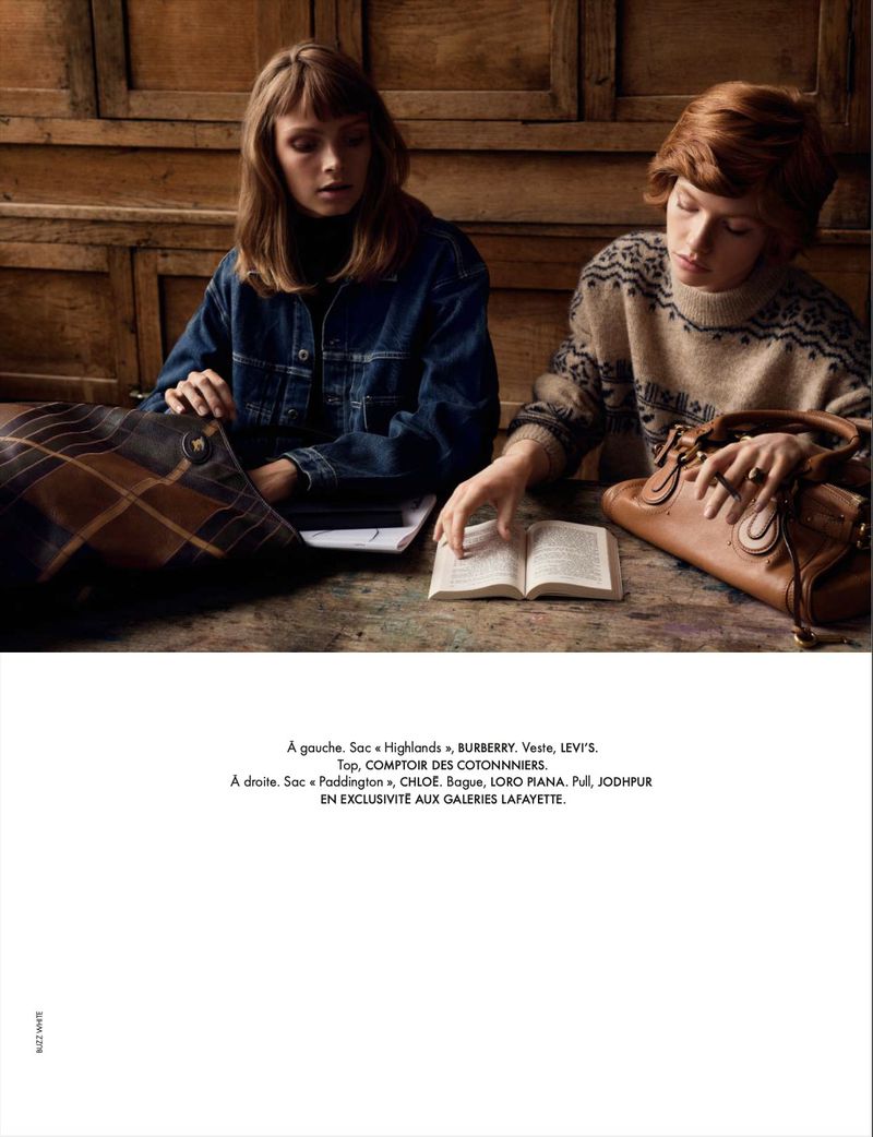 Two women sit at a wooden table with books and bags in a warm-toned indoor setting. One wears a denim jacket, the other a patterned sweater.