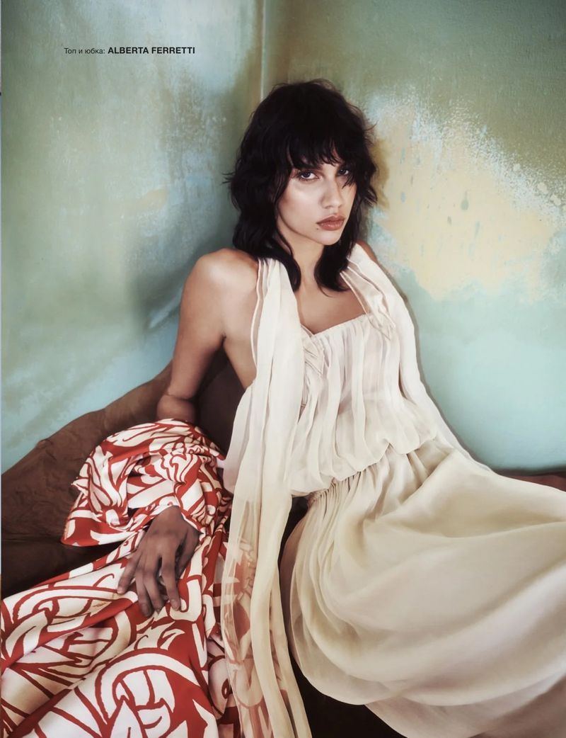 Woman in an off-white gown sits on a green and brown couch, posing for a fashion shot with a flowing gown and red patterned shawl.