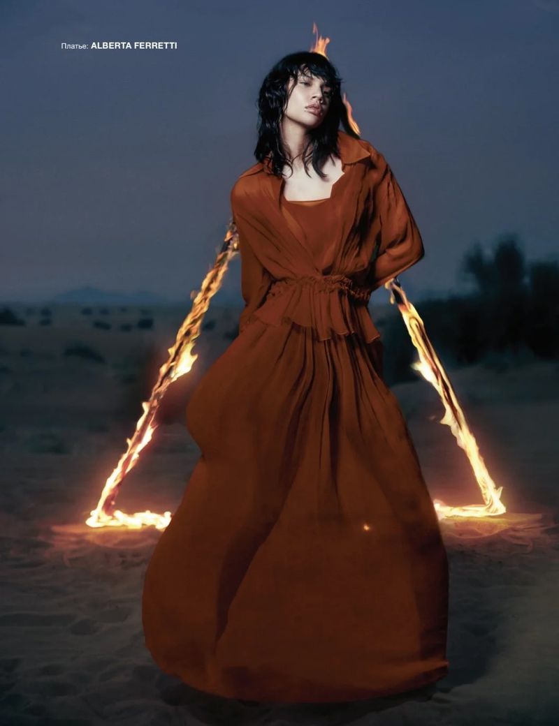 A woman in a brown dress poses in a desert setting with a triangular fire element behind her in a fashion editorial shot.