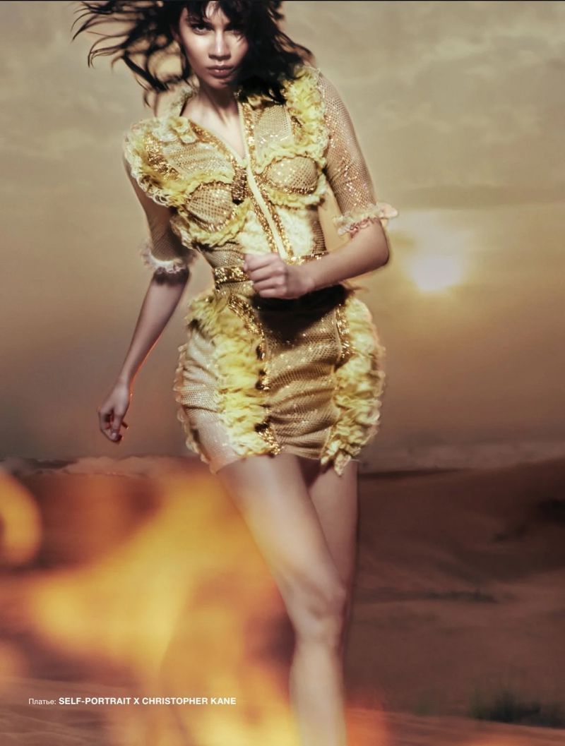 Fashion shot of a young woman in a golden embellished dress in motion, with a sunset sky background and orange glow.