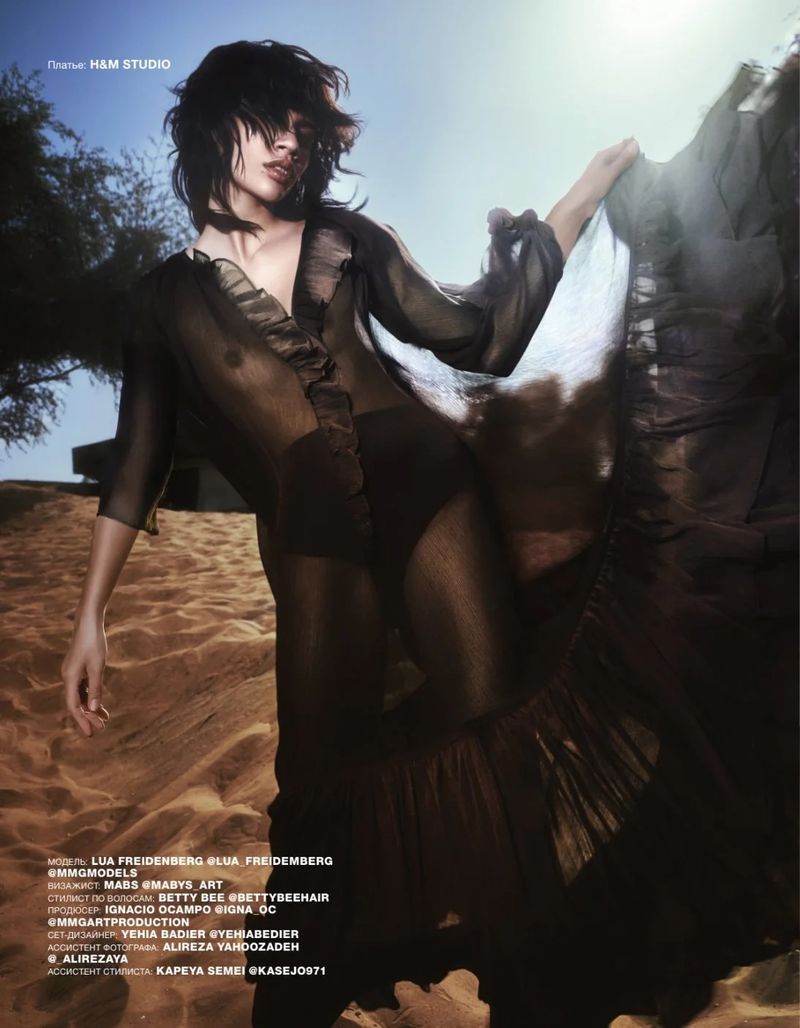A woman in a sheer dress poses in a desert landscape, illuminated by sun, creating a dramatic fashion shot for H&M Studio campaign.